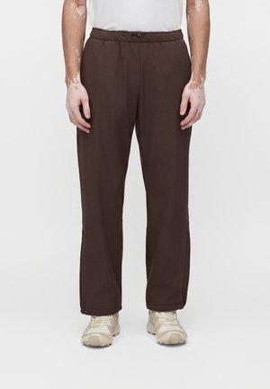 Persoană purtând pantaloni maro închis, cu croială lejeră și talie elastică, și adidași bej, stând în fața unui fundal alb simplu.