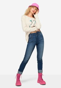 Volný krémový svetr s grafickým designem, modré džíny a jasně růžové kotníkové boty. Modelka má také růžový klobouk s širokým okrajem. Uvolněný outfit.