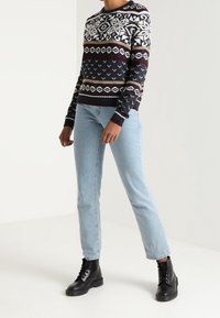 Pull en maille bleu marine avec des motifs géométriques blancs et bordeaux, assorti à un jean bleu clair à jambes droites et des bottines noires.