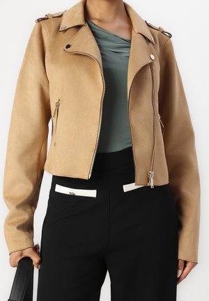 Femme portant une veste courte beige, un haut vert drapé, un pantalon noir avec des accents blancs aux poches, tenant un sac à main noir à ses côtés.