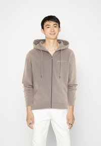 Sudadera con cremallera marrón en tejido suave, con un corte relajado, bolsillos tipo canguro y capucha con cordón. Presenta un discreto detalle del logo en el pecho.