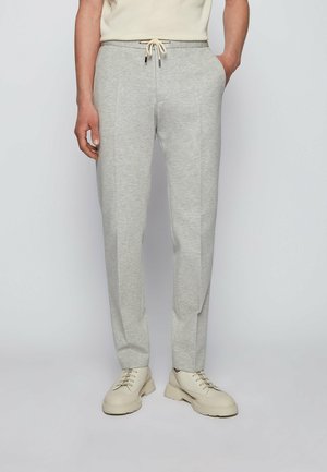 Mężczyzna w jasnoszarych spodniach z sznurkiem i kremowo-białych tenisówkach, stojący na jednolitym jasnym tle.