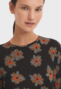Woman with tied-back hair wearing a black shirt with large abstract flowers in brown, gray, and red tones on a white background.