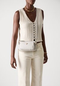 Hellgraue Leder-Crossbody-Tasche mit goldenen Beschlägen, strukturiertem Oberflächenfinish und Logodetail. Kombiniert mit einem cremefarbenen Strickoberteil und hellen Hosen.