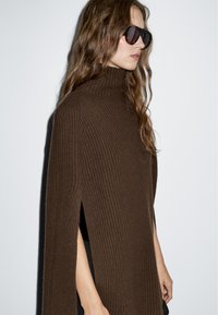 Brązowy sweter typu golf z prążkowaną fakturą i bocznymi rozci�ęciami. Modelka nosi czarne, oversize'owe okulary przeciwsłoneczne. Gładka tekstura na jednolitym tle.