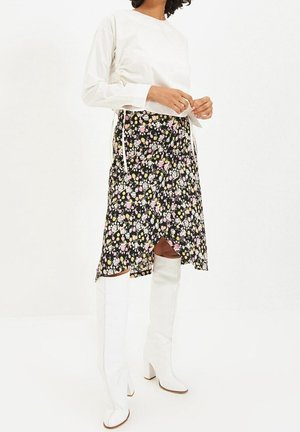 Frau trägt ein weißes langärmeliges Oberteil, einen schwarzen Midirock mit Blumenmuster und weiße kniehohe Stiefel mit Absatz, steht vor einem schlichten weißen Hintergrund.