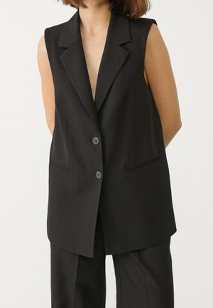 Woman wearing a sleeveless black tailored vest with lapels and two buttons, paired with matching black trousers, standing against white background.
