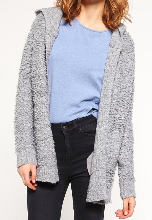 Femme portant un t-shirt bleu clair rentré dans un jean noir et un cardigan gris texturé, ouvert sur le devant avec une capuche.