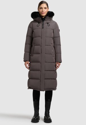 Cappotto imbottito lungo e grigio con cappuccio in pelliccia sintetica nera, caratterizzato da cuciture verticali, due tasche laterali e un taglio dritto. Abbinato a stivali neri al ginocchio.