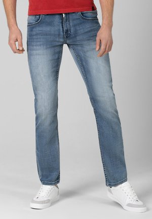 Mann trägt hellblaue ausgewaschene Jeans, weiße Turnschuhe und ein rotes Hemd und steht vor einem schlichten Hintergrund.