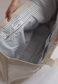 Bolsa de lona con exterior beige y forro de tela a rayas grises. Cuenta con un bolsillo con cremallera dorada y una etiqueta en el interior.