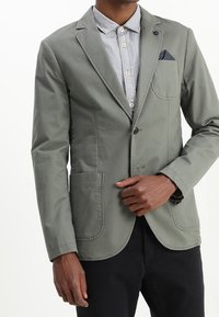 Mężczyzna w jasnoszarym blezerze z naszytymi kieszeniami, na jasnej koszuli z guzikami, z ręką blisko guzika blezera i w ciemnych spodniach.