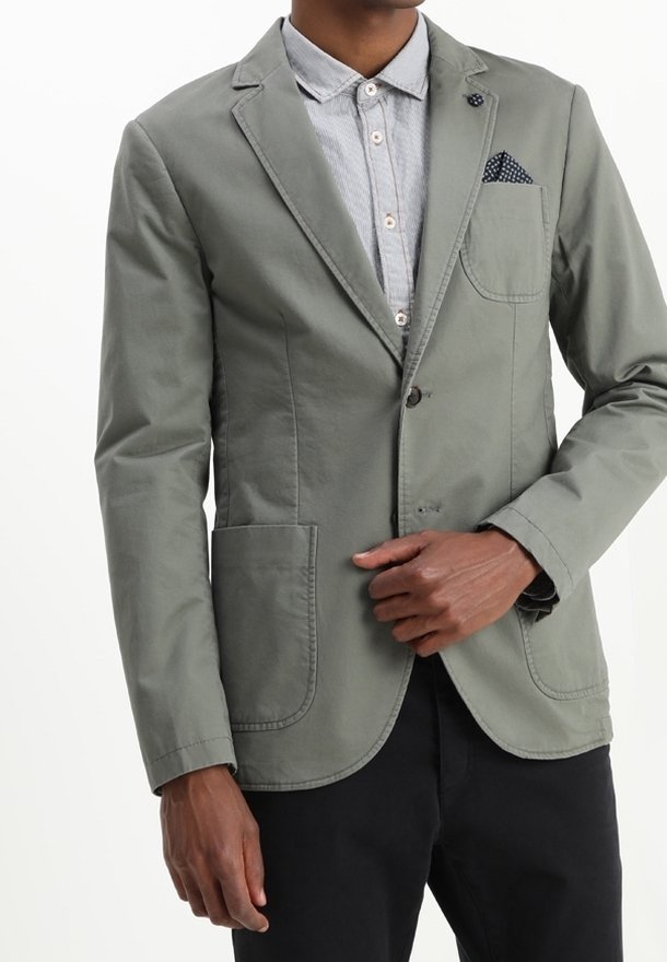 Mężczyzna w jasnoszarym blezerze z naszytymi kieszeniami, na jasnej koszuli z guzikami, z ręką blisko guzika blezera i w ciemnych spodniach.