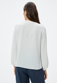 Witte blouse met lange mouwen en een subtiele textuur, ronde halslijn en uitlopende mouwmanchetten, gecombineerd met marineblauwe broek met streepjes.