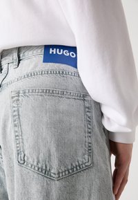 Svijetlosivi traperice s istaknutom plavom oznakom "HUGO" na struku. Imaju klasičan dizajn džepova. Mekana tekstura s suptilnim šavovima.