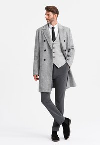 Abrigo gris de doble botonadura con grandes botones, superpuesto a un chaleco gris claro y pantalones gris oscuros. Botines de ante negros completan el conjunto.