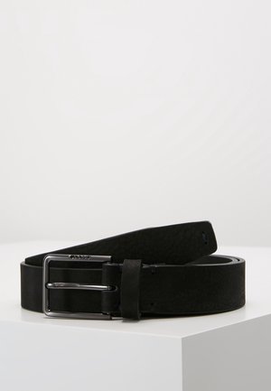 Ceinture en cuir noir avec boucle métallique rectangulaire argentée, enroulée et posée sur une surface blanche contre un fond clair uni.