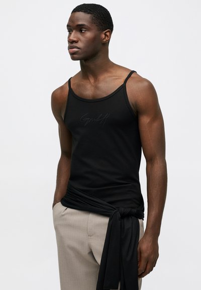 Noor mees mustas tank topis, millel on peene logo, beežides pükstes ja must riie, mis on tal vöö ümber kinni seotud, seisab ühtlasel taustal.