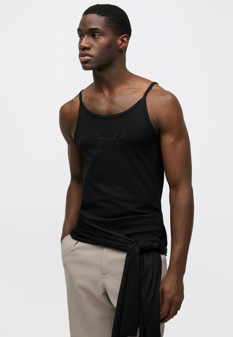 Giovane uomo che indossa una canotta nera con un logo discreto, pantaloni beige e un capo nero legato in vita, in piedi davanti a uno sfondo neutro.