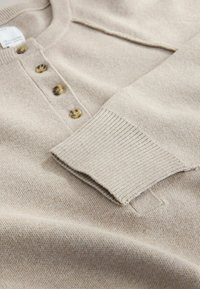 Beige neulepaita henley-kauluksella, jossa on kolme ruskeaa nappia ja resorihihansuut. Pehmeä tekstuuri ja rento muotoilu.