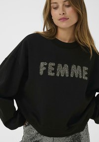 Naine kannab musta sviitrit, millel on hõbedast sädelevad rhinestone-kirjad "FEMME", koos sädeleva hõbedase seeliku ja vaatab allapoole.