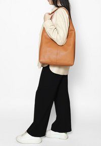 Bruin lederen tote bag met een textuurafwerking, met een ruime vorm en een enkele schouderband, vastgehouden door een persoon in neutrale kleding.