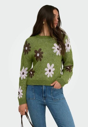 Kvinde iført grøn sweater med hvide og brune blomsterprint, blå jeans, holder en håndtaske, ser sidelæns med langt mørkt hår.