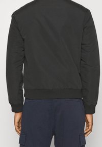 Veste bomber noire avec col et poignets côtelés, texture lisse et poches latérales, vue de l'arrière. Design léger et ajusté.