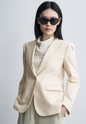 Γυναίκα με μακριά μαύρα μαλλιά που φοράει μεγάλα μαύρα γυαλιά ηλίου, εκρού σακάκι, ανοιχτόχρωμο πουλόβερ ζιβάγκο και παντελόνια σετ, ποζάροντας με τα χέρια στις τσέπες.