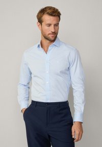 Lichtblauw button-up shirt van een gladde stof, lange mouwen en een klassieke kraag, gedragen met marineblauwe pantalon. Subtiele textuur, geen patronen.