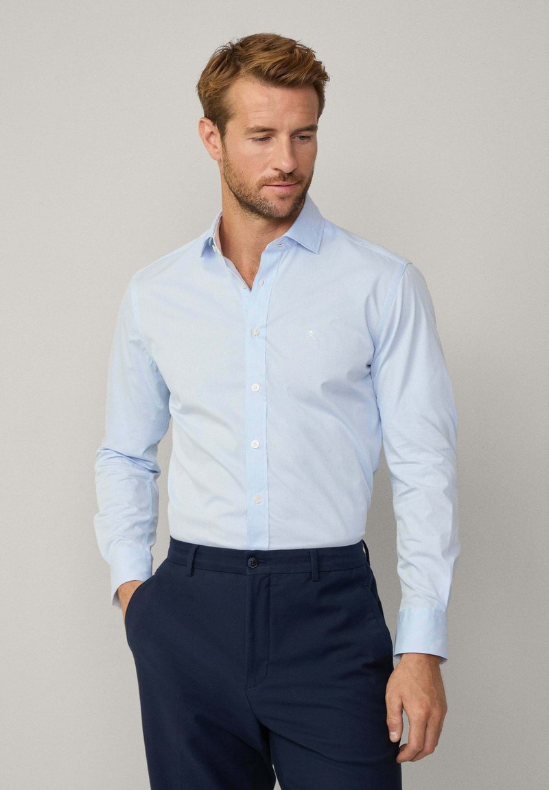 Lichtblauw button-up shirt van een gladde stof, lange mouwen en een klassieke kraag, gedragen met marineblauwe pantalon. Subtiele textuur, geen patronen.