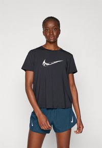 Svart T-skjorte med kort erm fra Nike med hvit logo, parret med marineblå shorts. T-skjorten har en avslappet passform og myk stofftekstur.