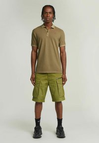 Polo verde oliva con maniche corte, abbinato a pantaloni cargo coordinati con più tasche e cerniera laterale. Sneakers nere e calzini.