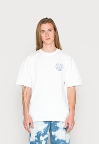 Hvit bomulls t-skjorte med løs passform og korte ermer, med en blå grafisk logo på venstre bryst. Kombinert med mønstrete blå denimjeans.