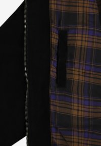 Czarna kurtka z zamkiem błyskawicznym z wewnętrzną podszewką w kratę w odcieniach brązu, czerni i fioletu. Posiada czarną kieszeń oraz teksturowany materiał.