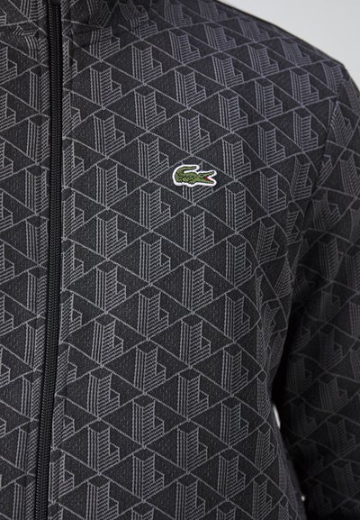 Veste à zip noire avec un motif géométrique, un tissu texturé et un petit logo crocodile brodé vert sur la poitrine.