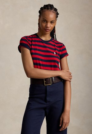 Femme aux cheveux tressés portant un t-shirt rayé rouge et bleu marine, un pantalon bleu marine et une ceinture marron foncé, debout, un bras croisé sur l'autre.