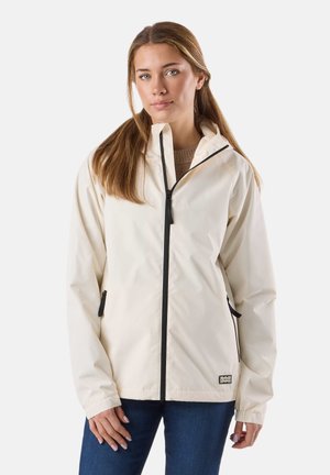 Jeune femme aux cheveux longs portant une veste crème zippée par-dessus un pull marron et un jean bleu, debout devant un fond blanc.