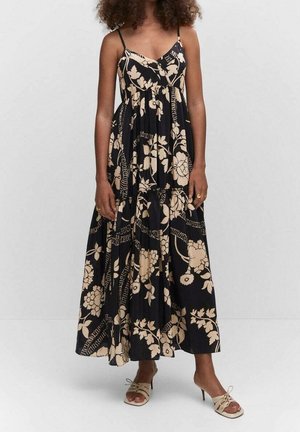 Femme portant une robe maxi noire à imprimé floral beige et bretelles fines, assortie à des sandales à talons ouvertes beiges.