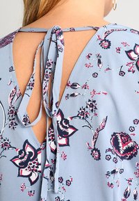 Tejido azul claro con un patrón floral en rojo y azul marino. Presenta un diseño de espalda en V con lazos y un ajuste relajado alrededor del escote.