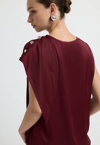 Top bordeaux con una texture liscia, caratterizzato da un design drappeggiato, maniche asimmetriche e bottoni metallici sulla spalla.