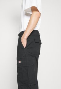 Vue de côté d'une personne portant un pantalon cargo noir avec des poches à rabat et une chemise blanche à manches courtes, la main dans la poche du pantalon sur un fond uni.