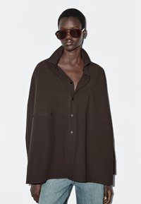 Camicia marrone oversize in un tessuto morbido con colletto alto, chiusura con bottoni e tasche laterali, abbinata a occhiali da sole scuri e jeans azzurri chiari.