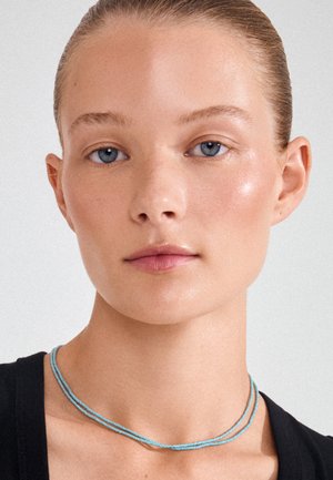 Ung kvinde med blå øjne og tilbagestrøget hår iført en dobbelt række lysblå perlekrans og en sort top mod en ensfarvet baggrund.