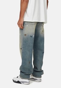 Lichtblauwe denim jeans met een losse pasvorm, voorzien van verfspetters en een vervaagde uitstraling. Gecombineerd met witte sneakers.