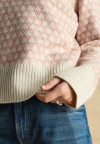 Hand, die einen silbernen Ring trägt, steckt in der Tasche einer blauen Jeans, und trägt einen cremefarbenen Pullover mit rosa geometrischem Muster und gerippten Bündchen.