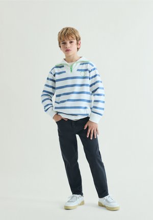 Băiat stând cu o mână în buzunar, purtând un pulover cu dungi albastre și albe, pantaloni închiși la culoare și adidași albi pe un fundal simplu.