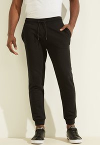 Zwarte joggingsbroek van zacht materiaal met een elastische tailleband, trekkoord, zijzakken en taps toelopende benen, gecombineerd met zwarte sneakers.