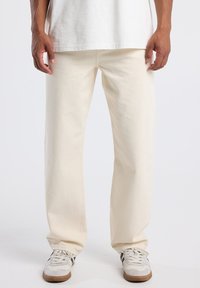 Hombre vestido con pantalones rectos color blanco roto, zapatillas blancas con rayas negras y una camiseta blanca, de pie frente a un fondo liso.