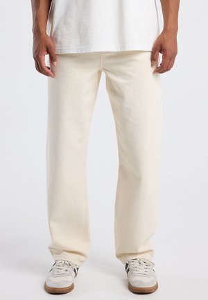 Uomo che indossa pantaloni dritti di colore avorio, sneakers bianche con strisce nere e una t-shirt bianca, in piedi contro uno sfondo neutro.